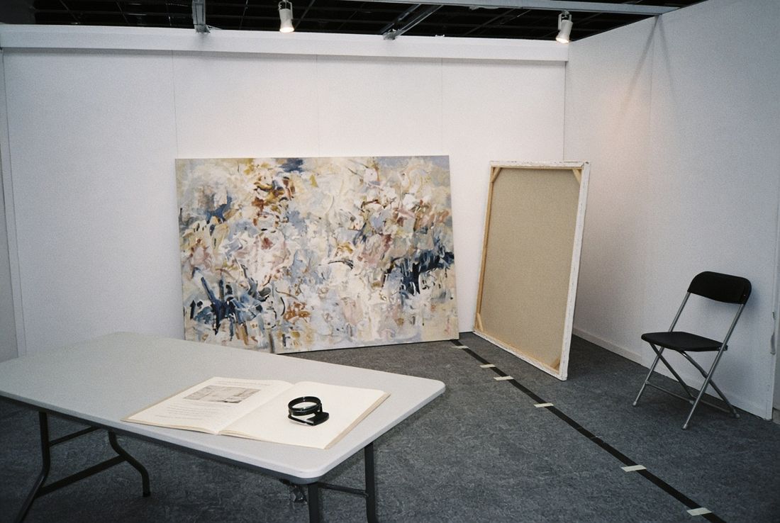 An art fair booth before opening, with a painting against the wall, a canvas turned face to the wall, and an open catalogue and loupe on a folding table